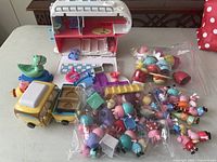 Photo showing scattered Peppa Pig figures in plastic bags, two yellow vehicles, and folded open pink and white camper playset on table.