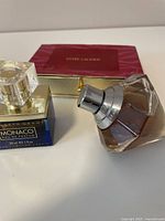 Photo showing Chopard Wish perfume bottle with clear faceted bottle, silver spray top, golden liquid, next to Elizabeth Grant Monaco Eau de Parfum square blue bottle with gold cap; Estée Lauder red and gold box in background.