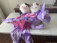 Purple plush dragon costume with colorful wings and tail laid flat on a table next to two cow-themed Squishmallows shows condition and details.