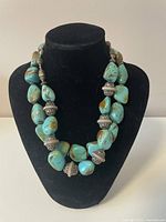 Photo of a chunky necklace with large turquoise stones and multiple ornate metal beads displayed on a black necklace bust.