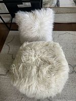 Photo shows a large round white faux fur pouf in front, and a large square white faux fur pillow behind, both on a rug.