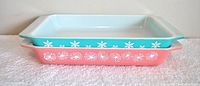 Two Pyrex pans stacked; pink floral design on bottom and blue snowflake design on top, both rectangular with integrated handles.
