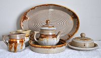 Full set arranged on white surface showing all 8 pieces including teapot, milk and water pitchers, covered butter dish, round dessert plate, and large oval platter. Pieces feature beige and brown drip glaze pottery design.