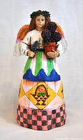 Front view of a hand-painted ceramic angel figurine with detailed colorful skirt and holding a flower pot with purple berries.