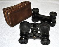Both pairs of black antique French opera glasses positioned on a white surface with the brown leather case visible beside them, showing size and condition