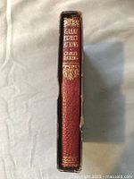 Side spine view of the antique red leather book with gold lettered title and author name, showing some wear around edges.