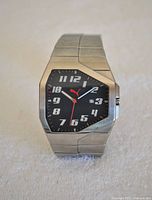 Front view of the PUMA watch showing its black asymmetrical octagonal dial, silver hour markers, and red Puma logo second hand with a date aperture.