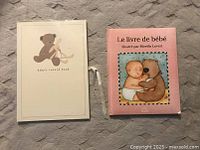 Two hardcover baby books side by side on a gray carpet: a white baby’s record book with a teddy bear and bunny illustration and a pink 'Le livre de bébé' illustrated by Mireille Levert with a baby and bear illustration.