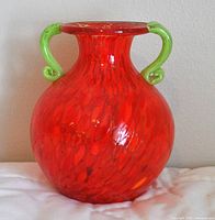 Front view of a bright red handblown glass vase with two green glass handles and rounded shape