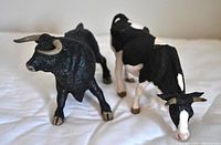 Black bull and black and white cow Papo figurines on white surface, showing frontal and side views