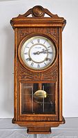 Front of wooden wall clock with carved decorative details, glass front panels revealing pendulum, and white dial with Roman numerals