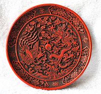 Front view of round Chinese carved lacquer plate showing detailed dragon and phoenix motifs surrounded by flowers in red and black.