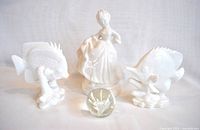 Photo showing all four items together: two white porcelain angel fish on each side, the Victorian shepherdess statuette at center back, and clear glass flower paperweight in front center.