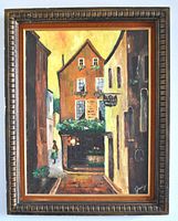 Full view of the framed oil painting showing a street scene with buildings, green plants, and a pathway. Signed on bottom right. The frame shows minor damage.