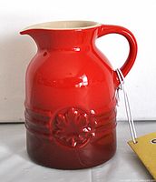 Front view of the red ceramic syrup jug showing embossed maple leaf design and handle with attached tag.
