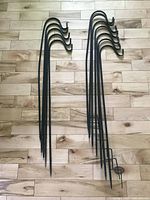 12 black metal shepherd's hooks arranged on floor showing top curves and foot ends, 7 with L-shaped anchors and 5 with straight ends.