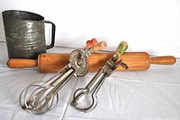 Four vintage kitchen utensils on white background: metal flour sifter, wooden rolling pin, two rotary mixers with wooden handles.