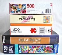 Stack of five 500 piece puzzle boxes showing titles and themes including Vintage Travel Tickets, Amazing Nature, Feline Frenzy, and colorful triangle pattern.