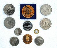 Top view of 15 commemorative tokens arranged on white background, showing various designs including 'The Dark Continent' brass token in original blue case, 1977 Silver Jubilee tokens with Elizabeth II on horseback, basketball and bobsleigh Olympic tokens, and several others.