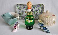 Seven decorative items arranged against neutral background including porcelain shoes, green glass cup, ceramic trinket box, Hummel figurine, blue and white pottery dish, and floral tray.