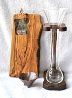 Front view of live edge olive wood cutting board with attached original tag, glass flower vase held in wooden stand, and small metallic pottery bowl.