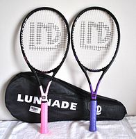 Pair of tennis rackets with one pink handle and one purple handle leaning against a black zippered carrying case showing the 'LUNNADE' logo on the case.