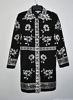 Front view of Nygard black coat with ecru thread floral embroidery, six covered snap buttons visible, collar and pocket placement shown.