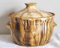 Front view of a round pottery covered bowl with artistic brushstroke glaze, showing the lid and side handles.
