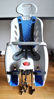 Front view of white molded plastic wrap-around bike child carrier seat with blue soft air cushion, three-point safety harness, and adjustable footrests showing mounting hardware beneath.