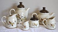 Front view of the full 7-piece Royal Doulton Lambethware tea and coffee set with Wild Cherry pattern showing water jug, coffee pot with lid, open cream jug, sugar bowl with lid, tea pot with lid, and milk jug lined up on white fabric.