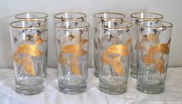 Front view of set of eight clear cylindrical drinking glasses decorated with large gold flying duck motifs and smaller black ducks.