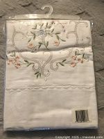 Folded white cotton pillow case in original plastic packaging with floral embroidery detail visible including pink and blue flowers with green leaves and small pearls.