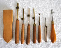 Top view of 8 vintage carpetmaking needles with wooden and metal handles and a wooden brush with fabric pad.