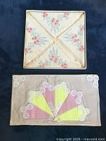 Top view of the floral paper napkins box with small pink flowers and greenery in folded arrangement and two fan-shaped pastel pink and yellow linen doilies edged in lace