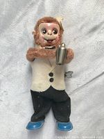 Full view of vintage wind-up monkey bartender figurine holding cocktail shaker while dressed in white jacket, black pants, and blue shoes.