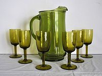 Full set of an olive green glass pitcher with six matching footed glasses displayed on a white surface against a white background.