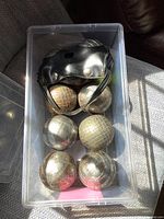 Top-down view of 8 steel chrome Petanque balls placed in a clear plastic storage container alongside a black leather carrying case with a shoulder strap.