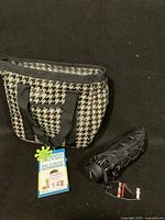 Photo showing houndstooth insulated lunch bag with black handles and a compact black folded umbrella with tags.