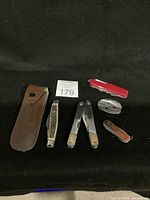 Five pocket cutters displayed on black surface along with brown leather sheath and auction lot number card