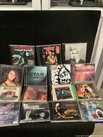 Photo of 16 visible CD cases showing album art by various artists in jewel cases, includes rock and pop genres.