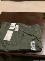 Folded olive green Perry Ellis pants with attached tags and multiple size 36 x 32 stickers, lying on a dark surface.