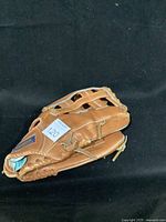 Front side view of tan leather softball glove folded with fingers together showing laced webbing and a paper number tag.