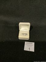 Front view of silver-colored ring with damaged clear center and side stones displayed inside white jewelry box on black background.