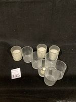 Photo shows 8 small clear glass candle holders placed on black fabric background. Three holders contain white candles, five are empty.