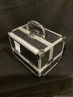 Closed rectangular black steel makeup container with silver edges and handle on black background.