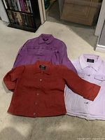 Three colored jean jackets laid flat on carpet showing purple, reddish-orange, and light lavender colors with front pockets and buttons visible.