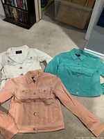 Three colored jean jackets (white, peach, turquoise) laid out on carpeted floor