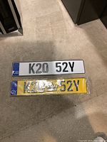 Top-down image showing both vehicle plates side by side on carpet floor. GB plate is white with embossed letters and blue EU/GB band, Cyprus plate is yellow with black letters, blue EU/CY band and crown symbol, sealed in plastic.