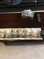 Panoramic black and white photo spread out on a table showing a large group of men seated and standing outside in front of a building.