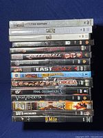 Photo shows a stacked view of 15 DVDs in shrink wrap and cases, titles visible including Saw, Exorcist, Trois 2, Rollerball, Constantine, Snoop Dogg Eastsidaz, Goldmember, Dogma, Final Destination 3, V for Vendetta, King of the Cage, South Central, and 8 Mile.